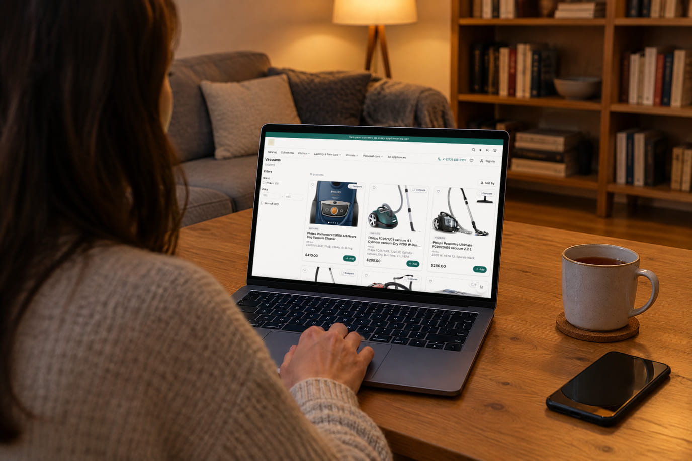 A shopper at home chooses a vacuum on a laptop: the screen shows the real online storefront with product cards and prices