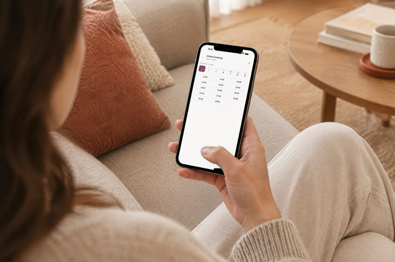 A client at home on the sofa books a master on her phone, with the salon’s free times on screen